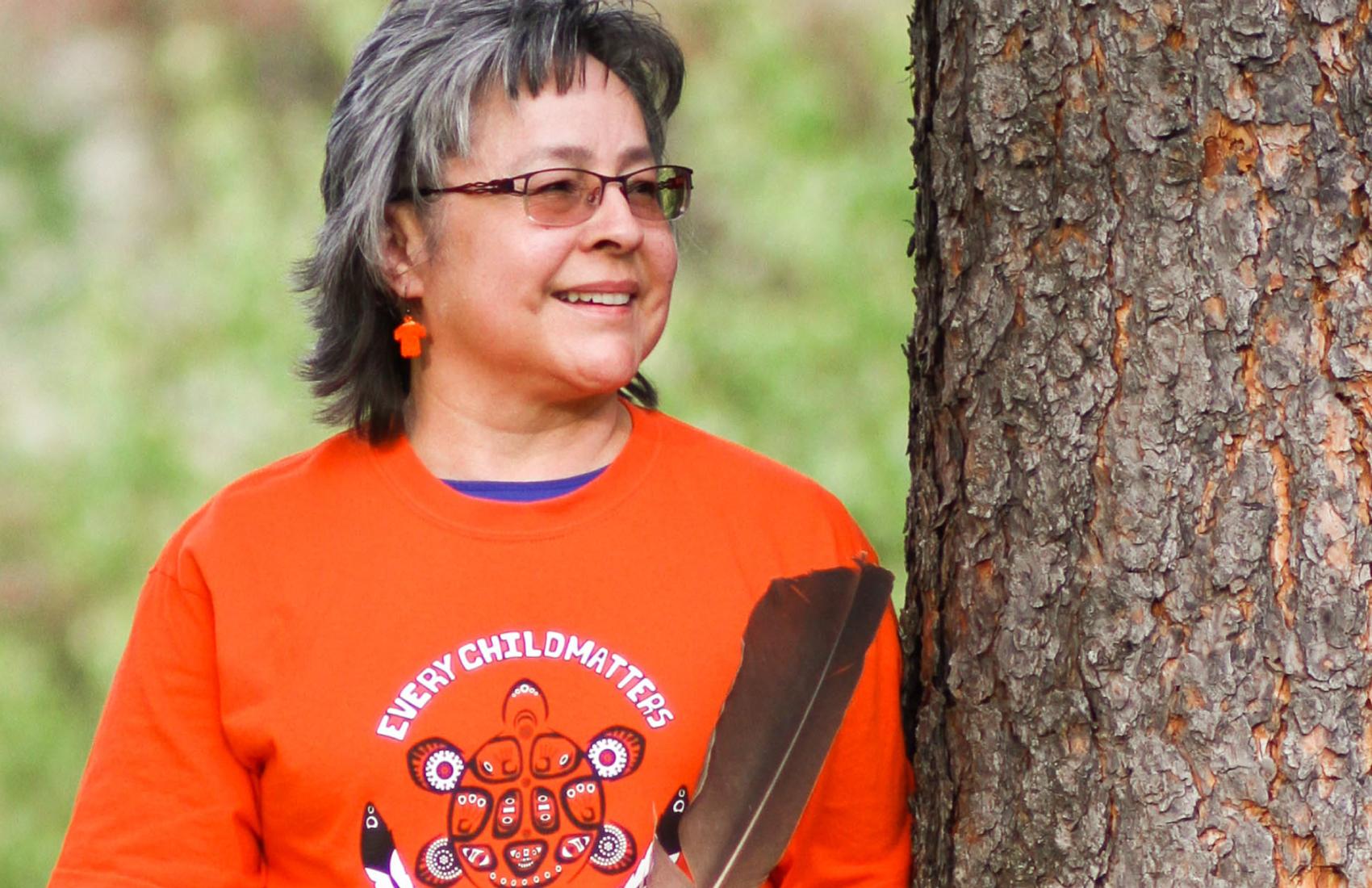 Phyllis Webstad, founder of Orange Shirt Day, stands next to a tree and holds a feather. She is wearing an orange shirt with the phrase "Every Child Matters"
