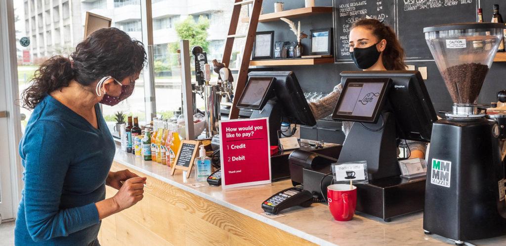 A patron wearing a COVID-19 masks tries to pay for purchase from mask-wearing cashier at a coffee shop