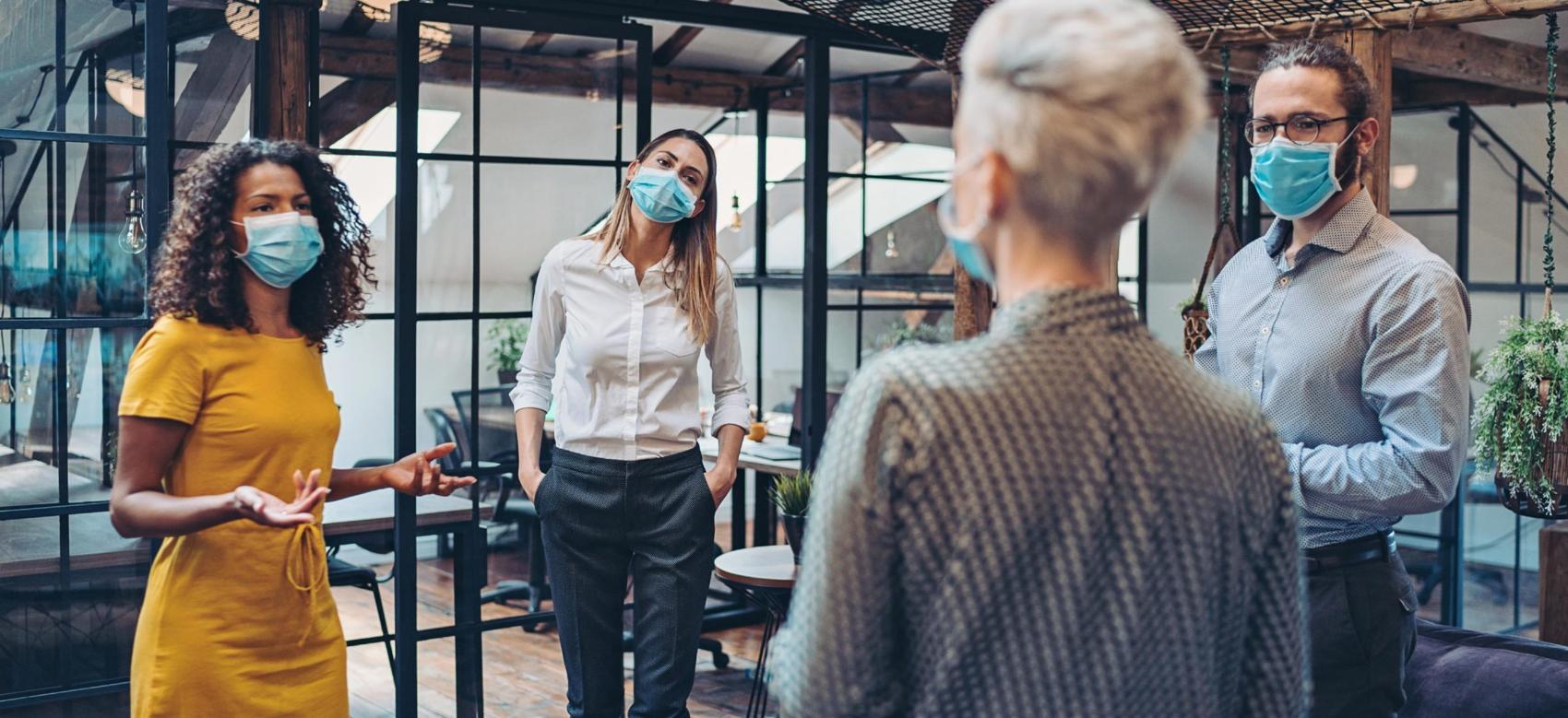 Four adults gather together in an office to discuss work in a post-COVID world. All are wearing medical masks and keeping distance from each other to adjust to re-opening.