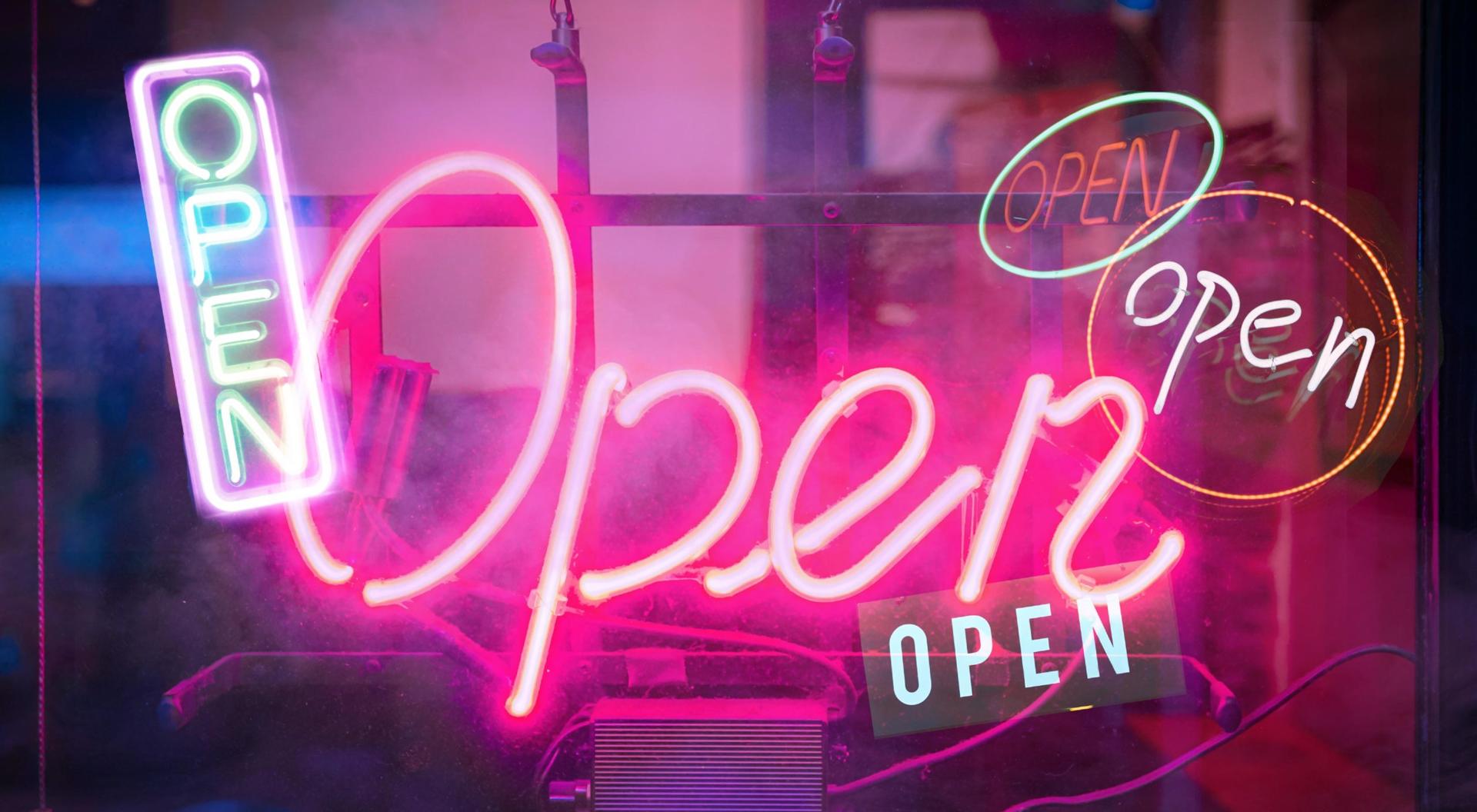 A photo illustration of several neon "Open" signs alight in the dark to signify re-opening of society after COVID-19