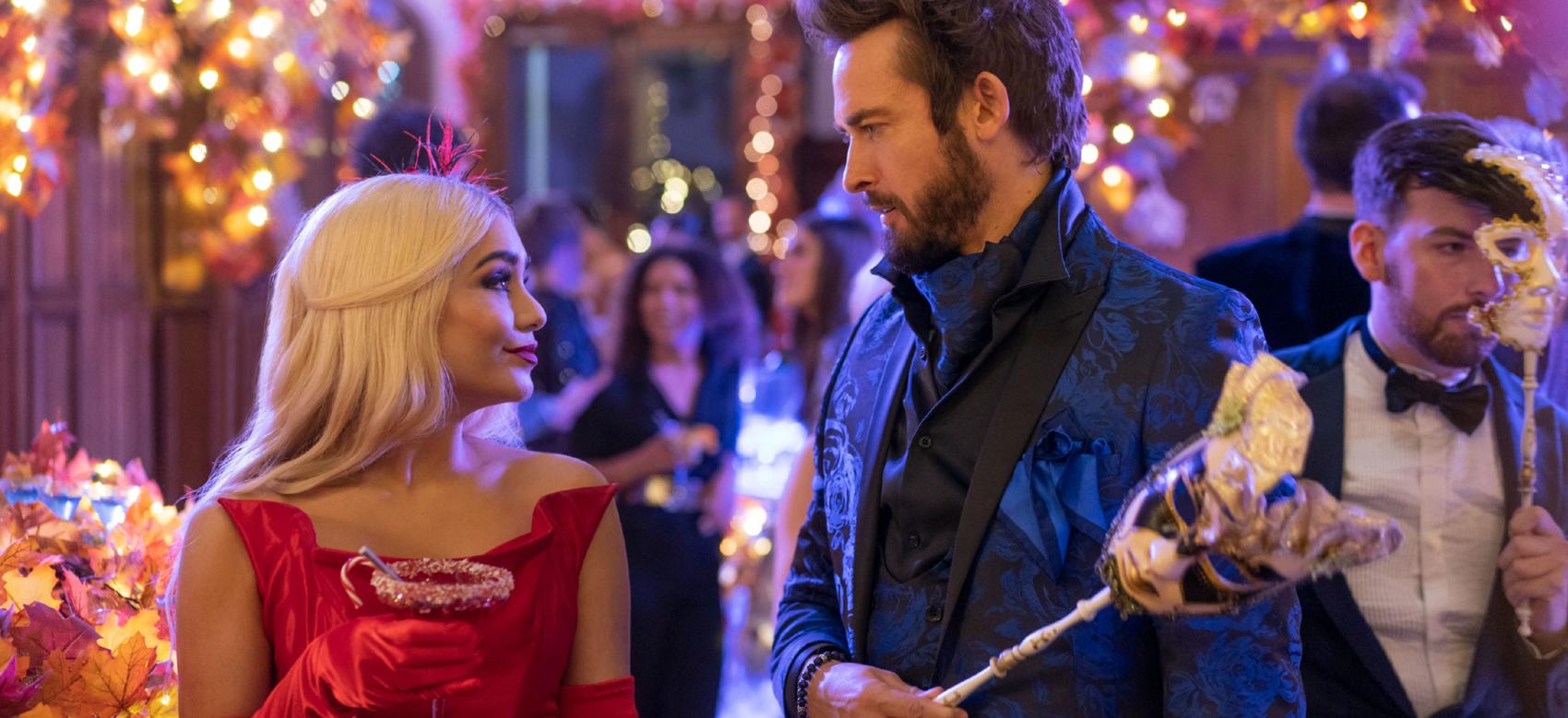 Woman in red dress holding cocktail and man in blue suit holding a mask at masquerade party surrounded by lights