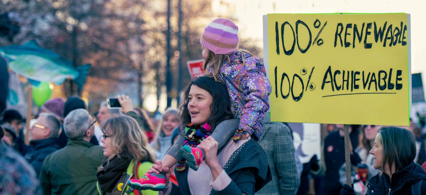 A group of people at a climate change protest. In the centre of the photo is a young woman with a child on her shoulders