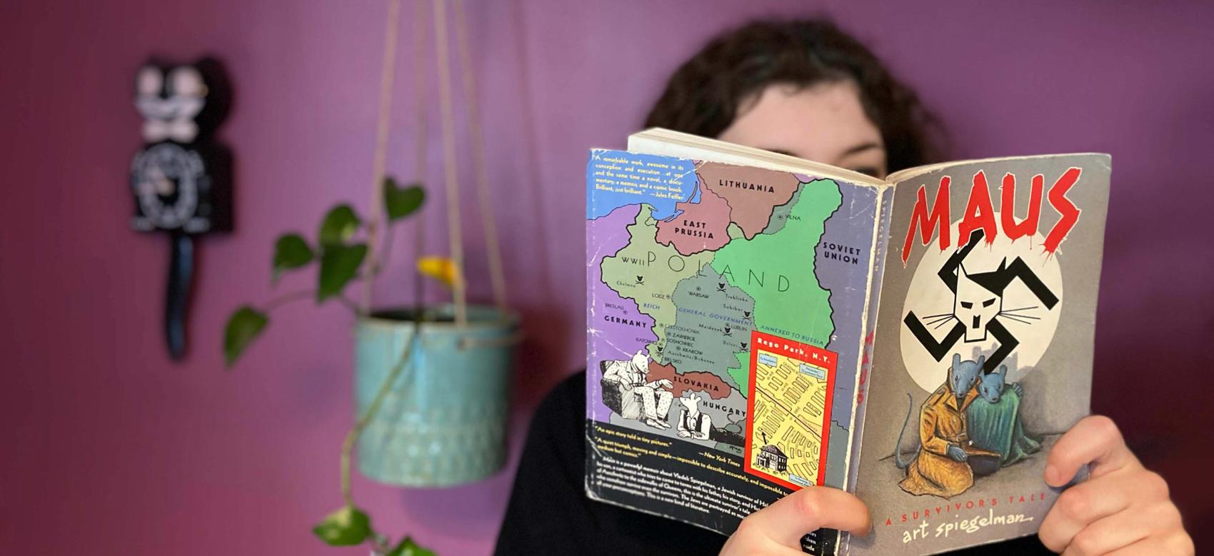 Photo of a young woman reading the book Maus with a colourful background with a plant and a cat clock over her shoulder