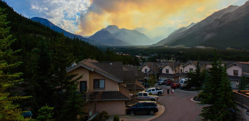 Wildfire smoke in the distance threatens a suburban community of large houses with SUVs parked in the driveways.