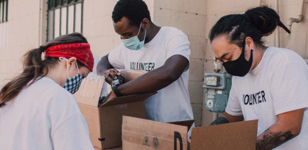Three people wearing shirts that say 'volunteer' pack boxes with food.