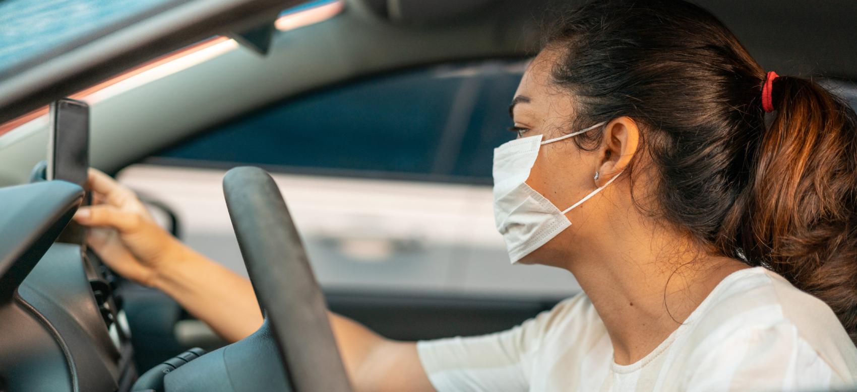 A woman wearing a mask in the driver's seat looking at a cellphone.