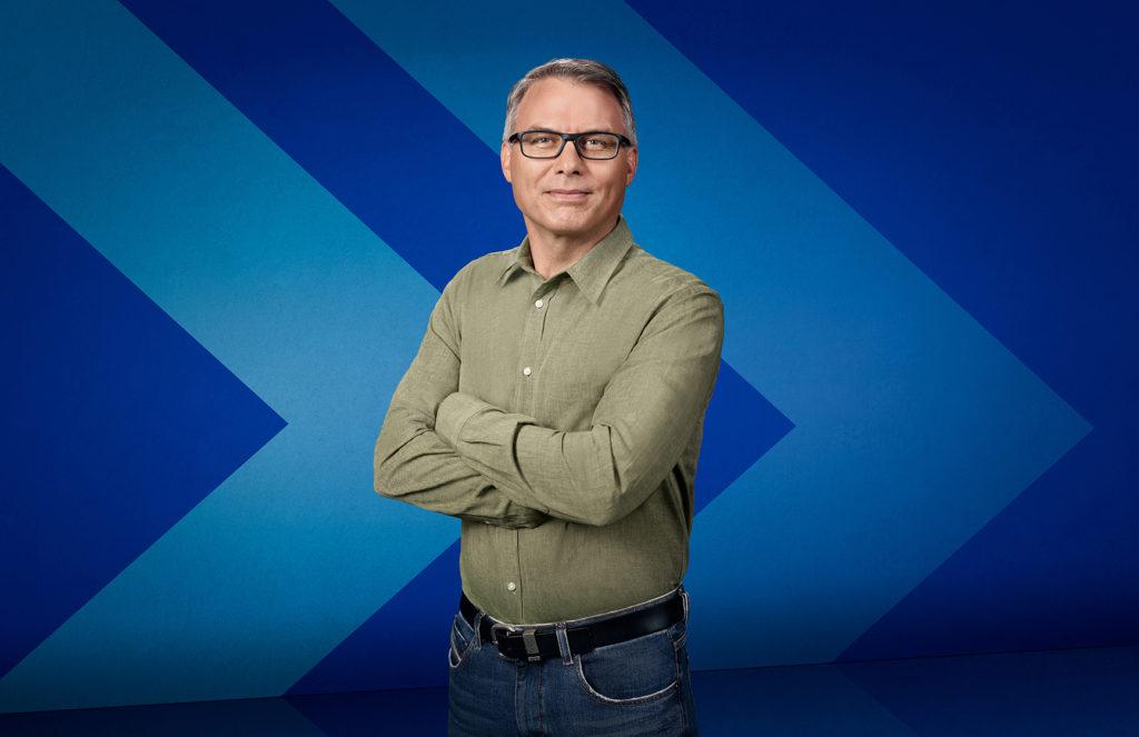 Dr. John Pawlovich, a family doctor who is the Rural Doctors’ UBC Chair on Rural Health, stands in front of a stylized chevron background