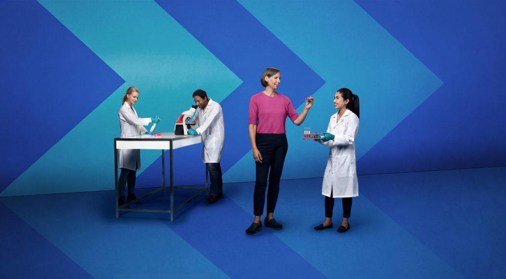 Three people in white lab coats and a person in a pink shirt working in a stylized lab with chevron backdrops.