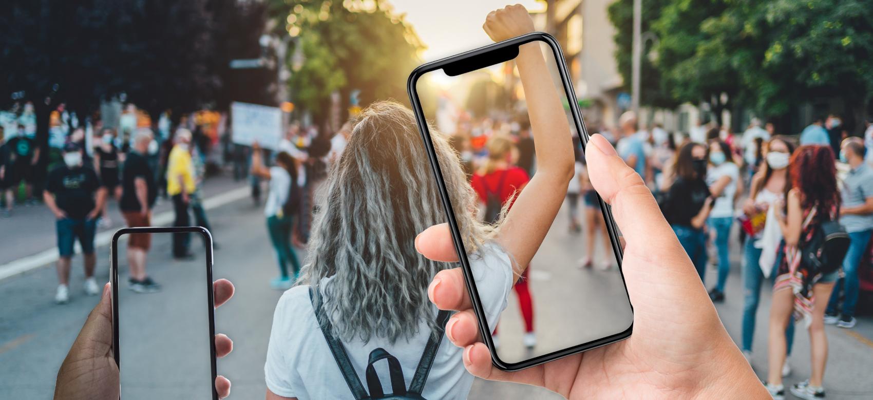 Hands holding up iPhones at a protest march in the streets, focused on the back of a person who has their fist in the air