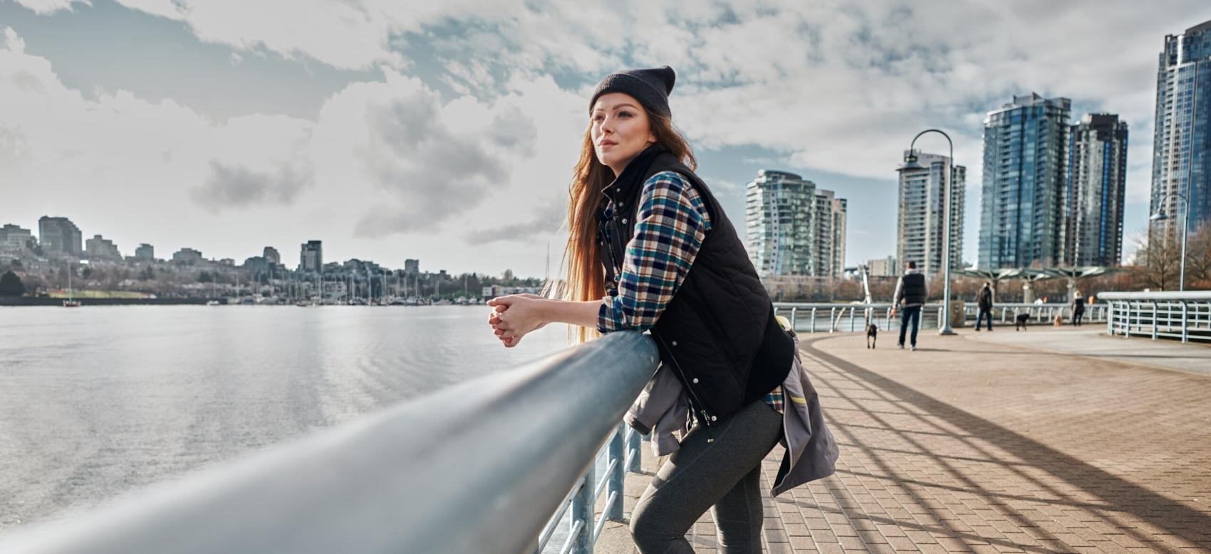 A lone person wearing a toque leans on the railing of the seawall in Yaletown, Vancouver, and looks out onto False Creek.