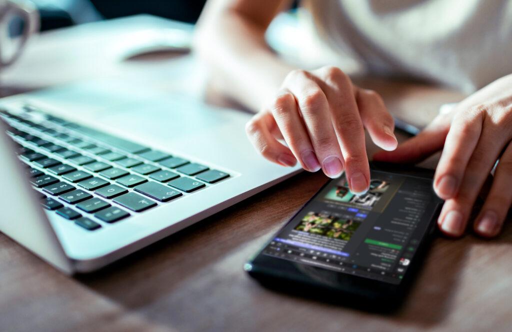 A person taps a phone next to an open laptop.