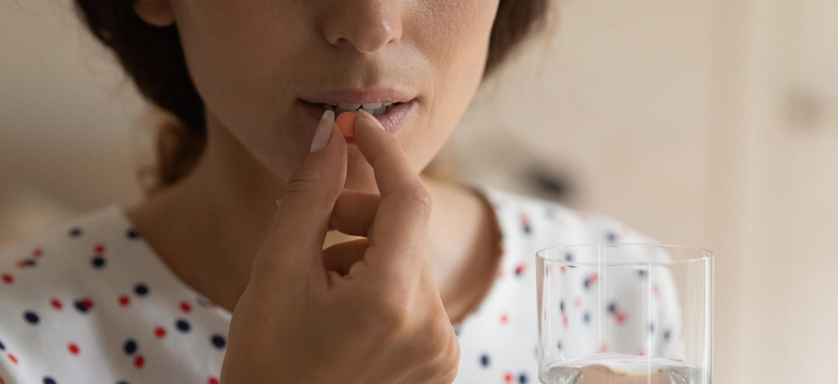 Close up cropped shot of young person holding a glass of water prepare to swallow an orange pill.