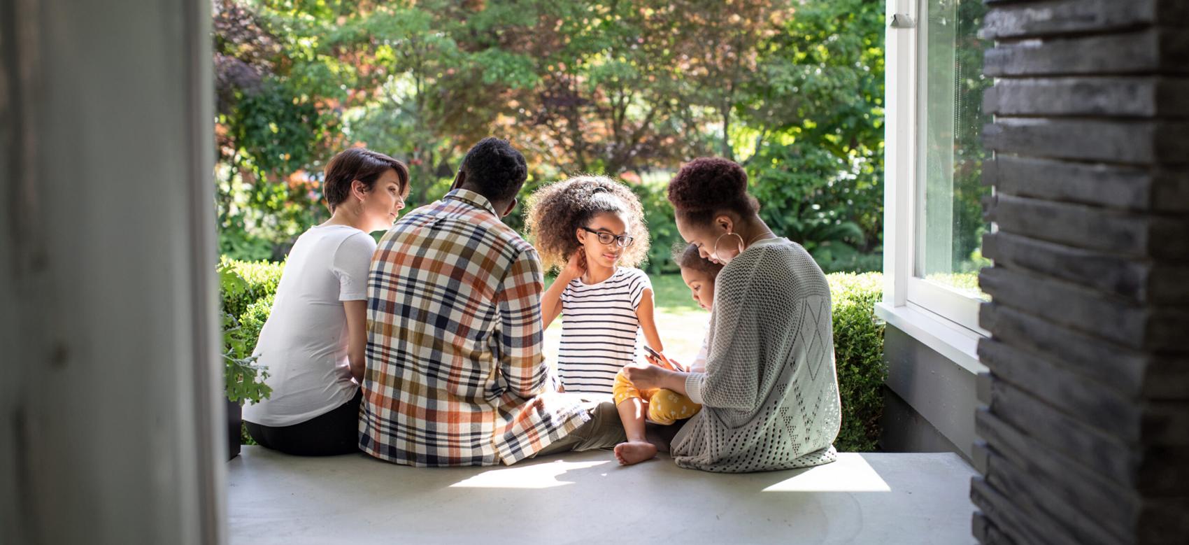 A Black family with a mother, father and three children sit beside one another on the porch.