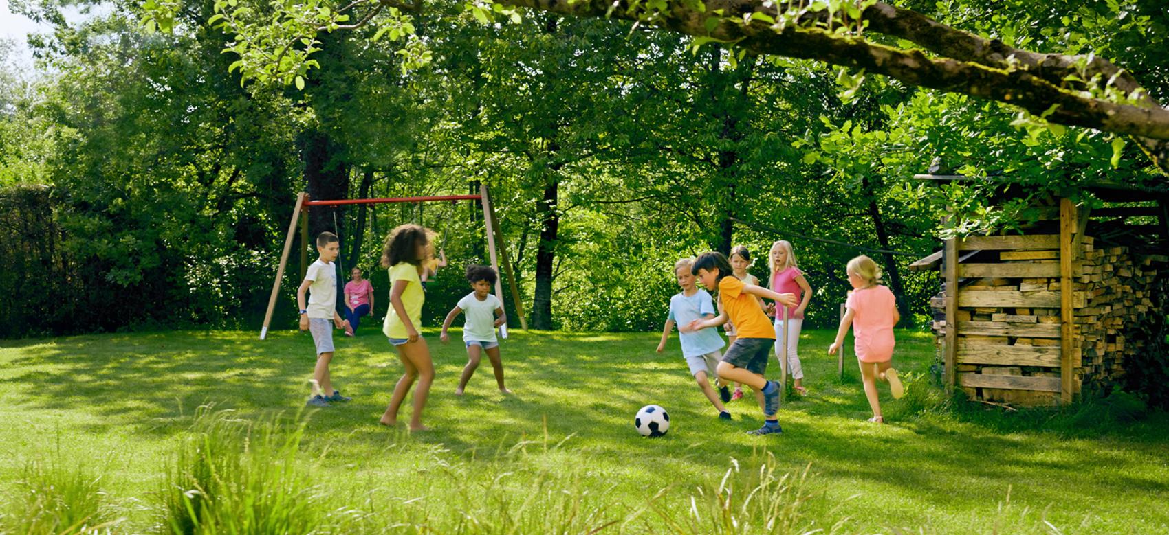 Children play soccer on grass outside with trees in the background and a swing set