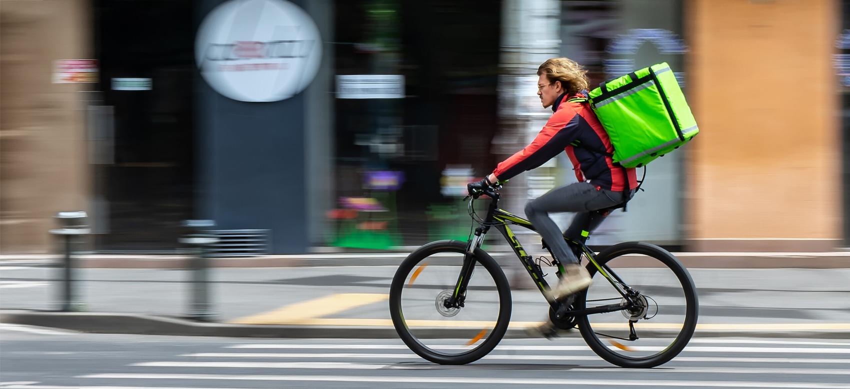 A cyclist wearing a red and black jacket rides quickly down a city street, carrying a large green delivery backpack, with blurred buildings in the background.