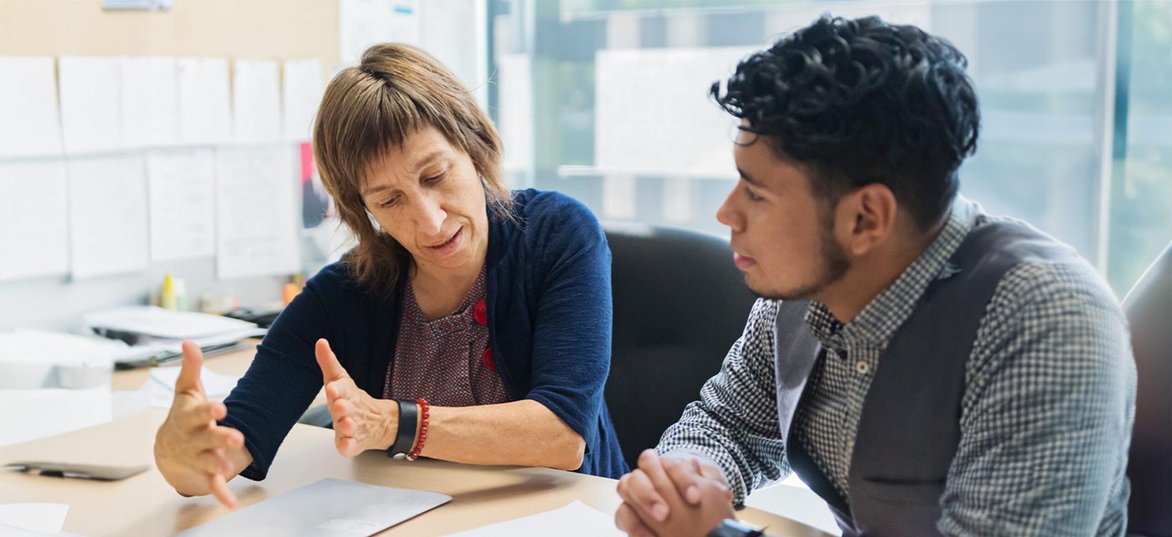 Two people sit at a desk at an office looking at paperwork and having an animated discussion.