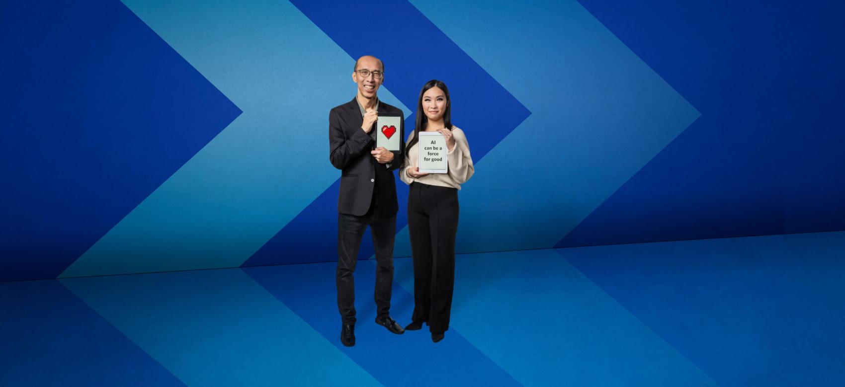 Two people stand smiling in front of a blue chevron background. Dr. Raymond Ng holds a digital printout of a heart, and Christy Kwok holds a sign that reads "AI can be a force for good".