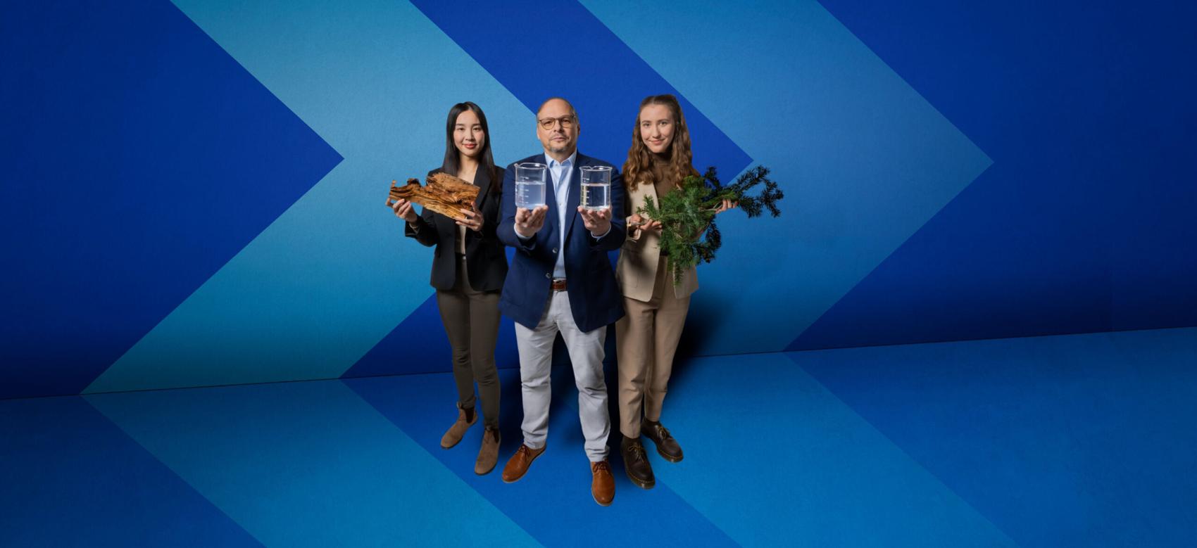 Three people stand in front of a blue chevron background, holding beakers of water, a tree branch and a piece of bark.
