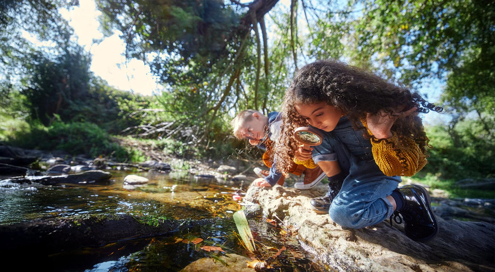 A boy and a girl looking at river with magnifier in the woods.