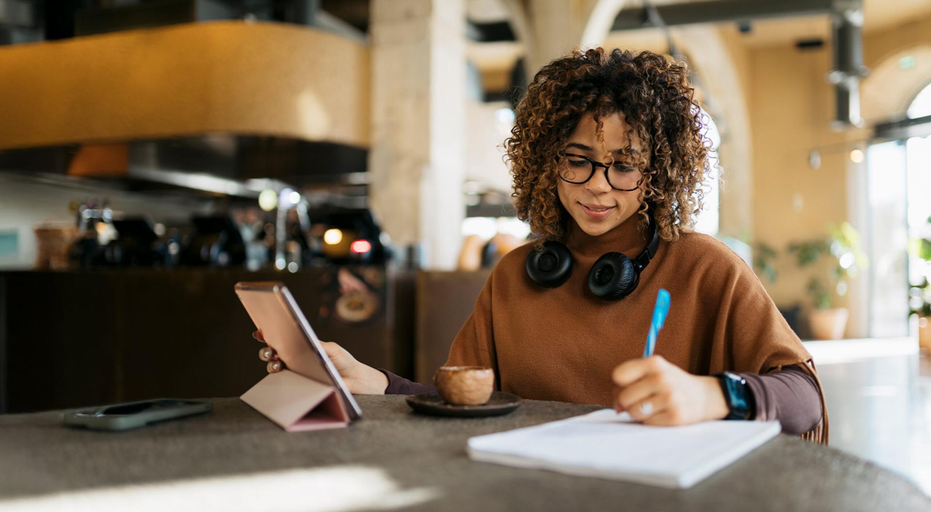 Young adult woman taking notes while using a tablet in a modern cafe setting.