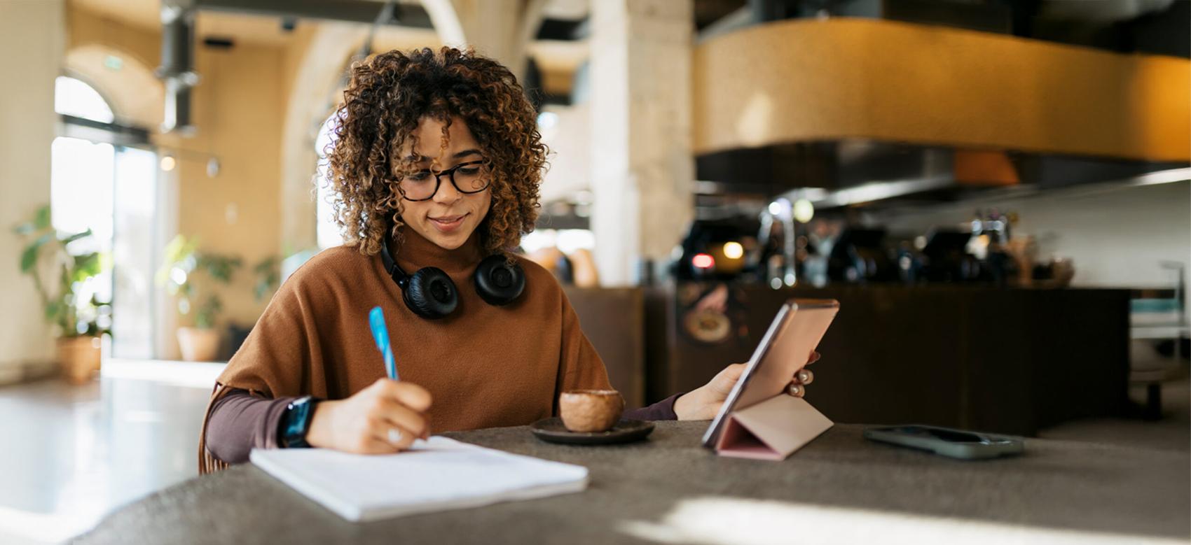 Young adult woman taking notes while using a tablet in a modern cafe setting.